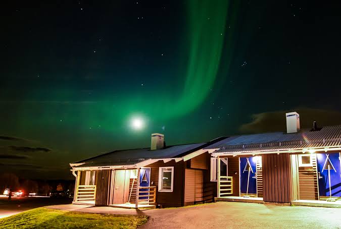 Low timber cabins at night with warm porch light under a green aurora.