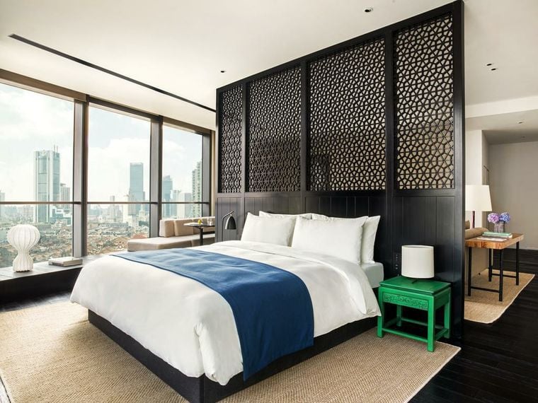 A hotel bedroom with a lattice screen behind the bed and a city view at the window.