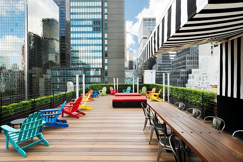A rooftop terrace with colourful chairs and a long table among city buildings.