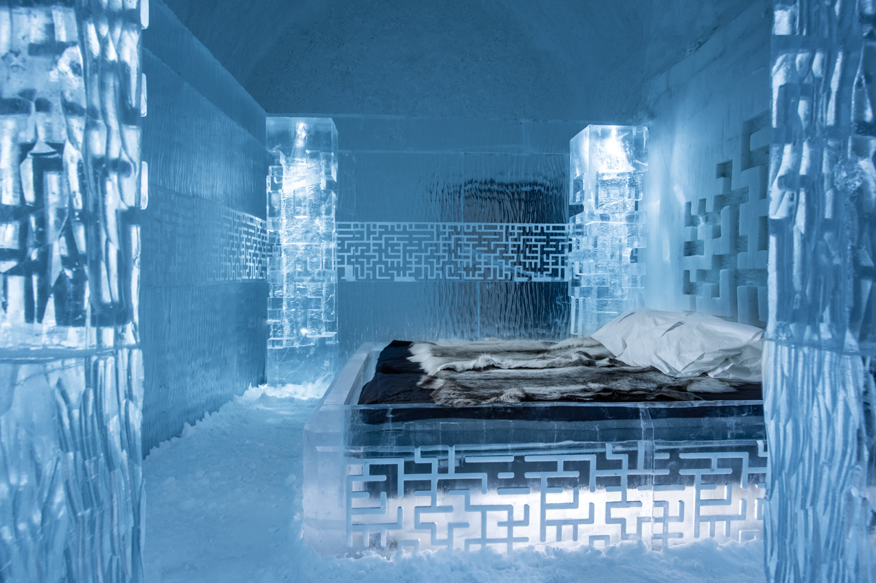 An ice-built hotel room with a carved ice bed dressed in furs under blue light.